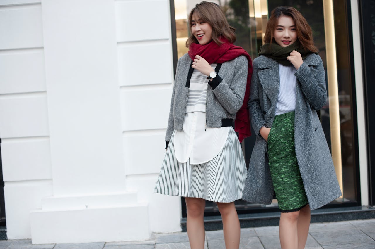 Two fashionable women wearing autumn clothing stand outdoors against an urban backdrop.