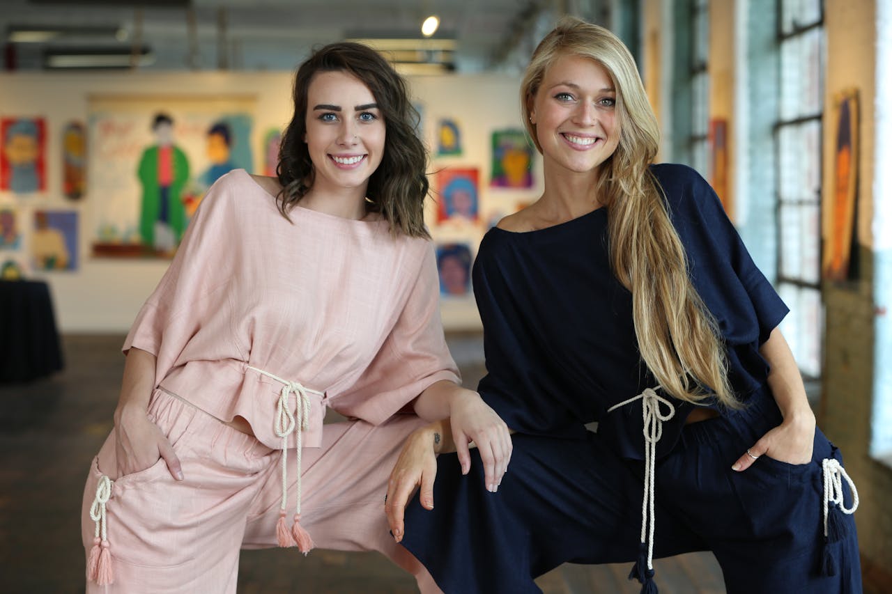 Two women smiling and posing indoors in fashionable outfits with an art gallery background.